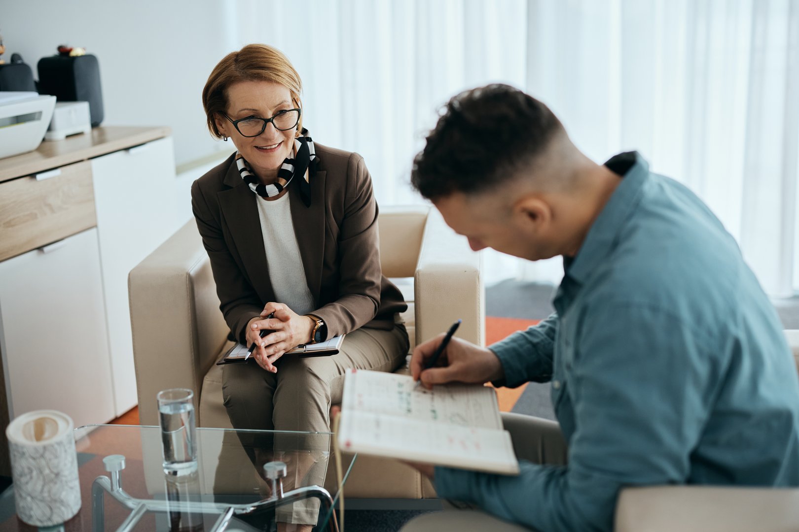 Happy mature psychotherapist talking to her patient who is taking notes during a counseling.