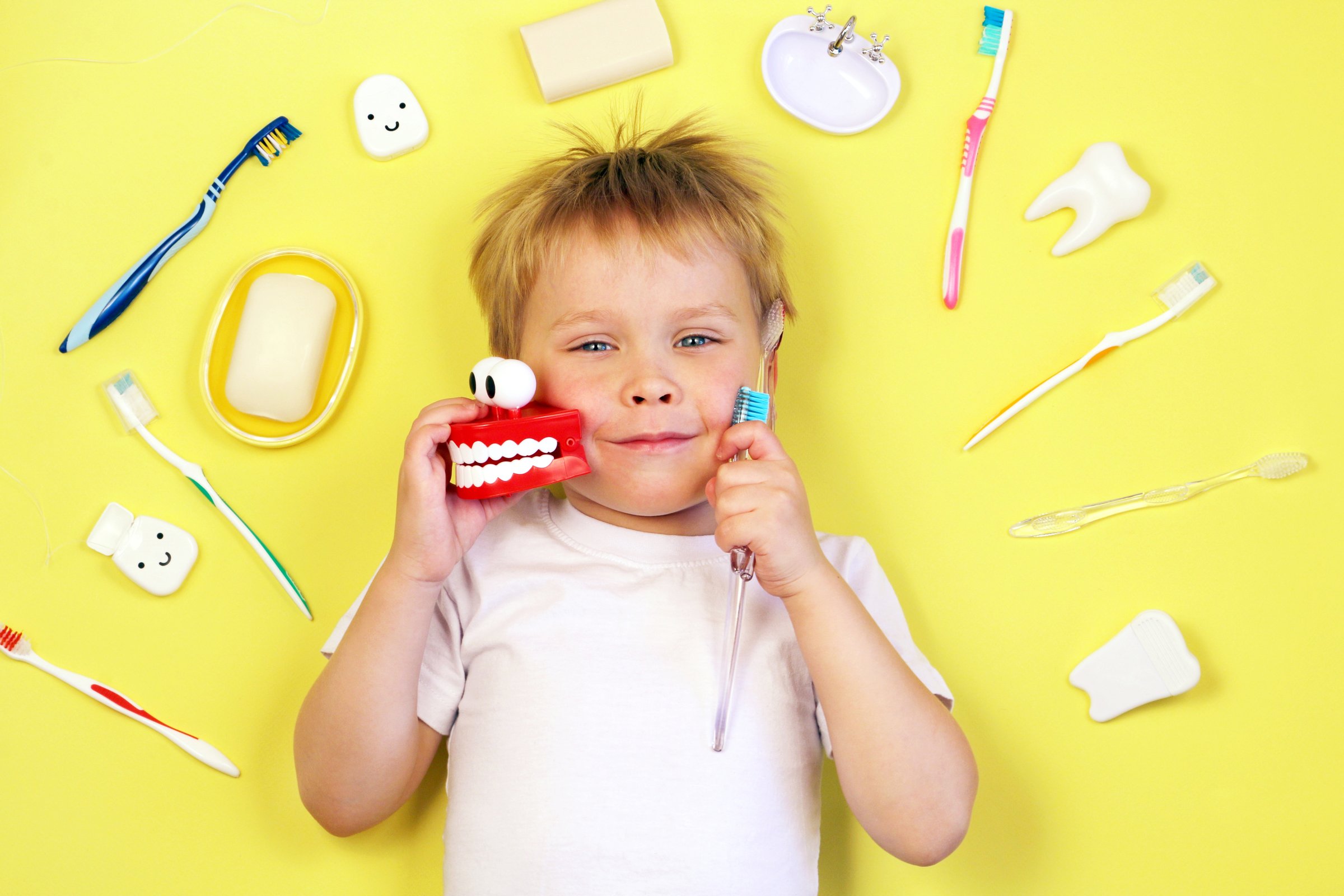 Cute boy child kid holding toothbrushes and toy teeth on yellow background. Teeth cleaning, oral care, dental hygiene concept , top view.