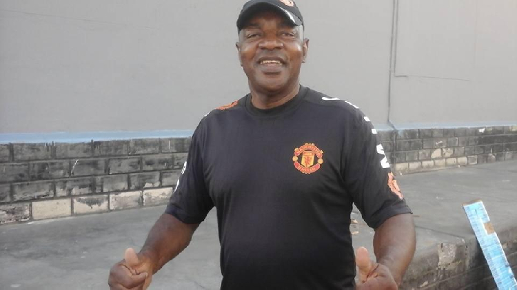 Smiling man in a Manchester United shirt with thumbs up, standing outdoors against a gray wall.