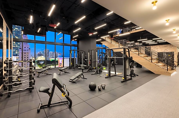 Modern gym with exercise equipment, including weights and benches, large windows, and a staircase leading to an upper level.