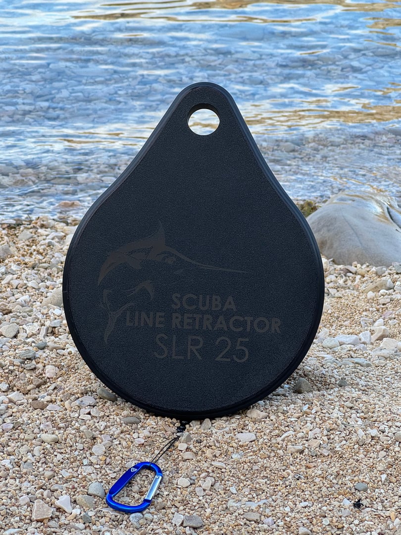 A Scuba Line Retractor SLR 25 on a pebble beach with a blue carabiner clip attached, near the waters edge.