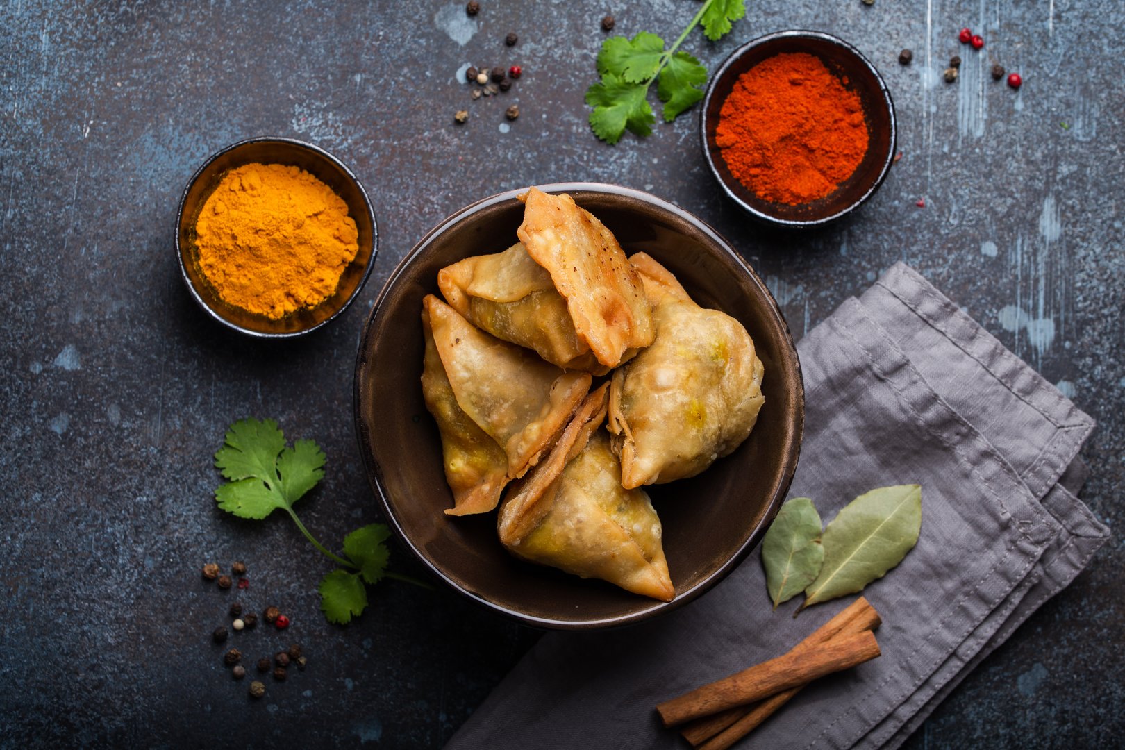 Indian samosas - fried/baked pastry with savoury filling, popular Indian snacks, served in bowl with spices and fresh cilantro on rustic background, top view. Overhead of samosas