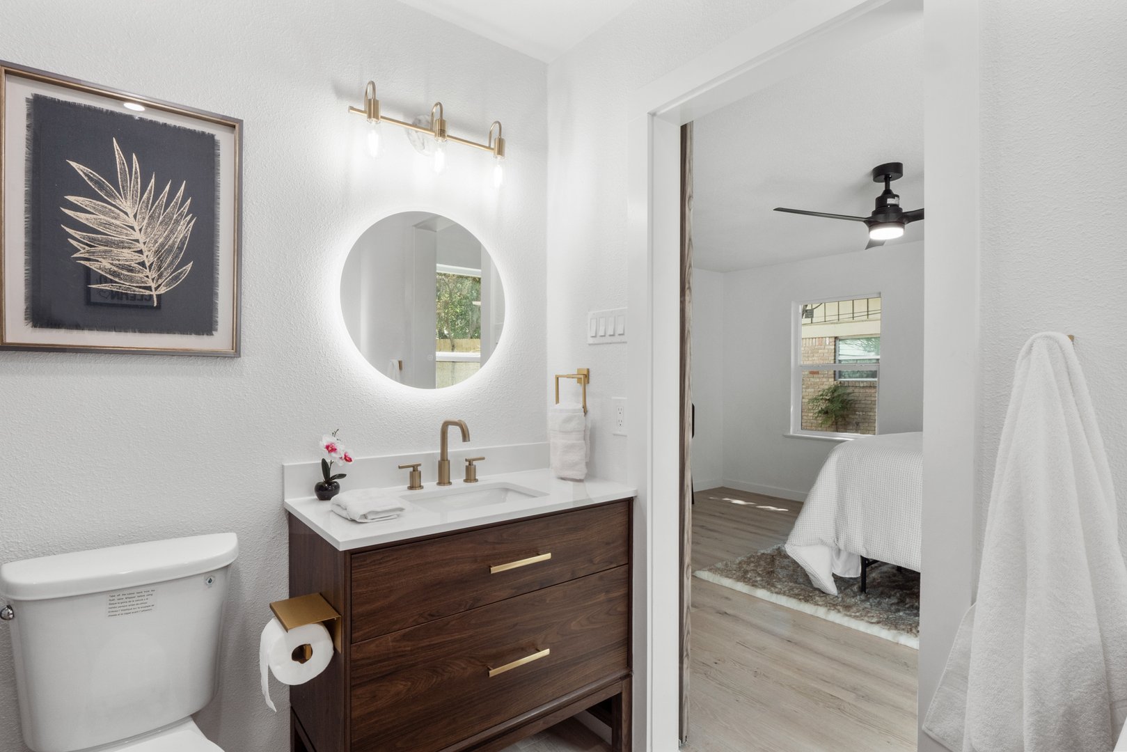 Modern bathroom with circular mirror, vanity with sink, toilet, and doorway leading to a bedroom with a bed and ceiling fan.