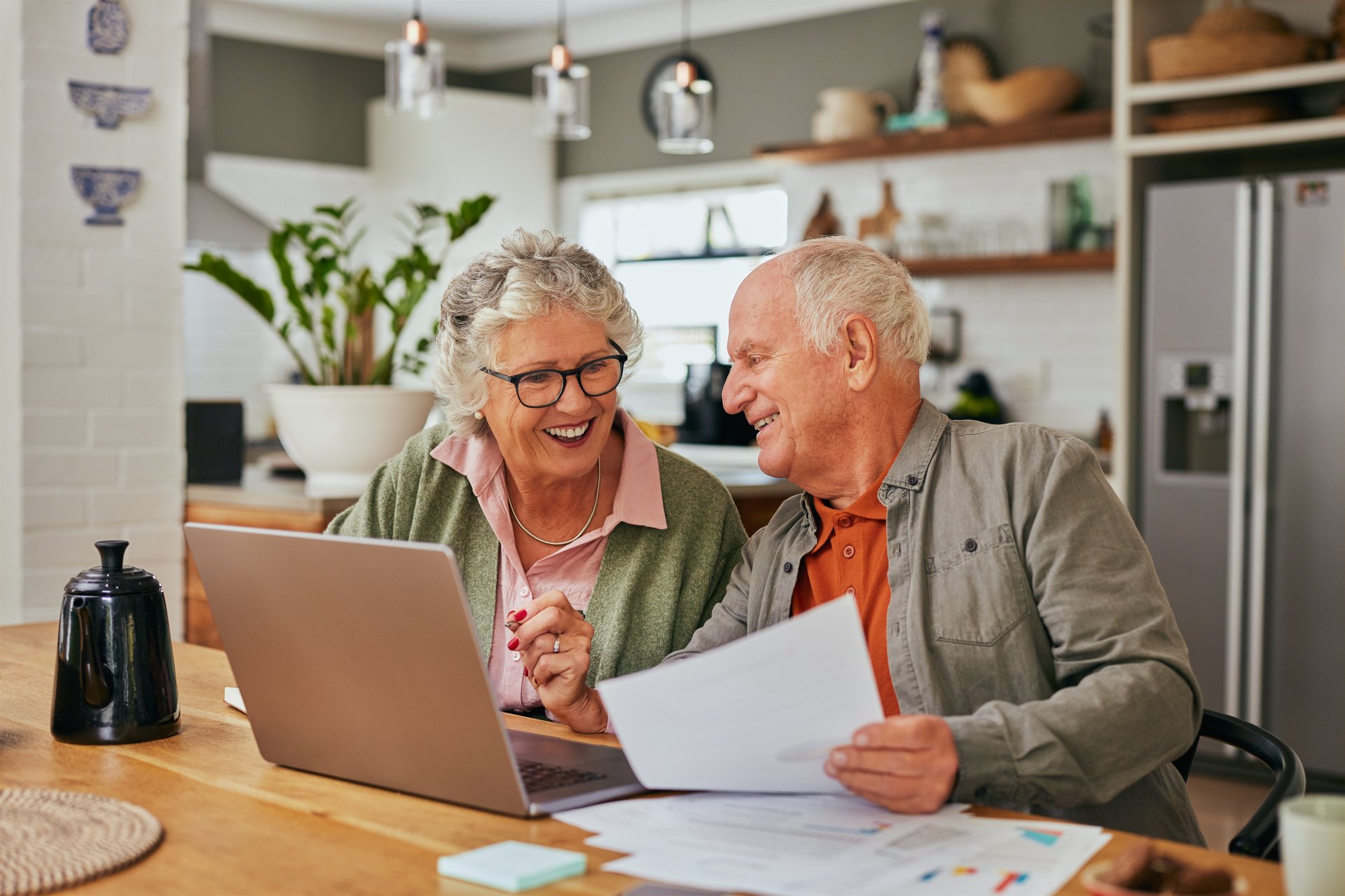 Smiling senior couple discussing bills and reviewing documents while using laptop at home. Mature couple collaborating on financial management at their dining table. Elder man and retired woman sharing a moment while organizing their finances while using computer.