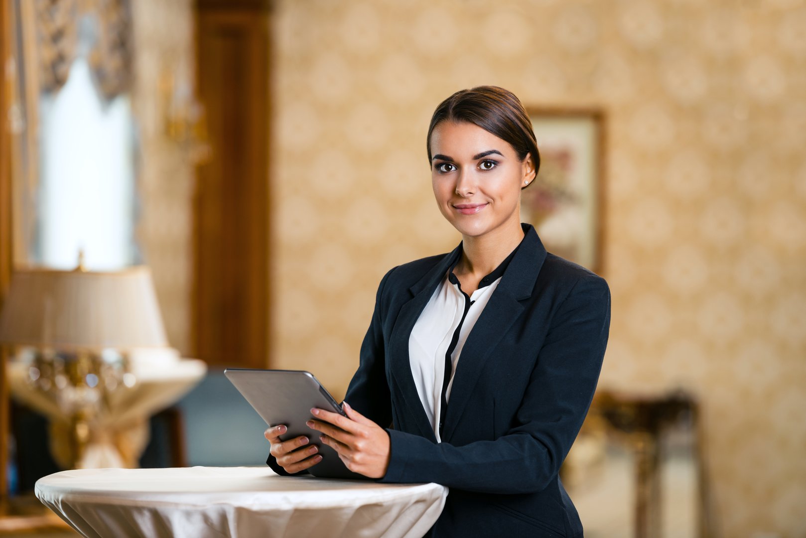 Photo of business woman in expensive hotel. Young business woman wearing suit, standing in nice hotel room, using tablet computer and looking at camera