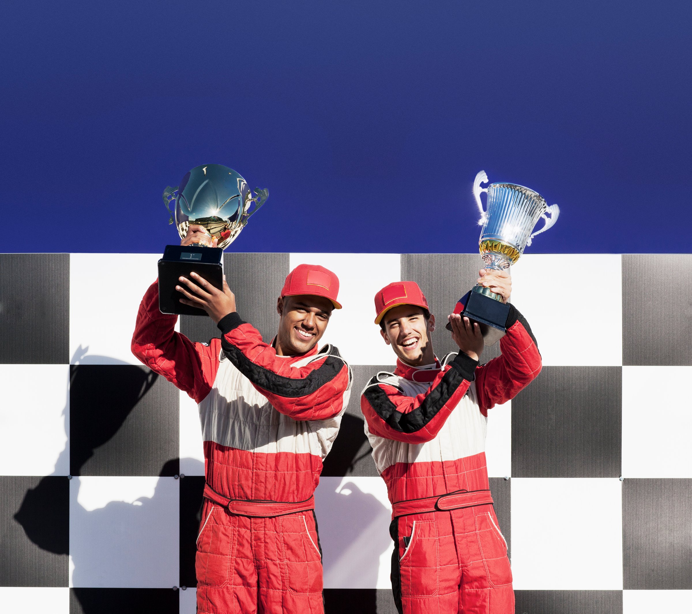 Two race car drivers in red suits and caps celebrate with trophies on a checkered podium against a clear blue sky.