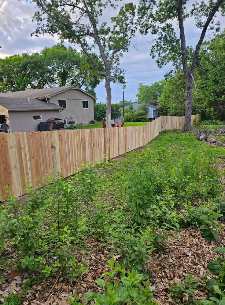 New wooden fence separates a grassy yard from a house with trees in the background under a cloudy sky.