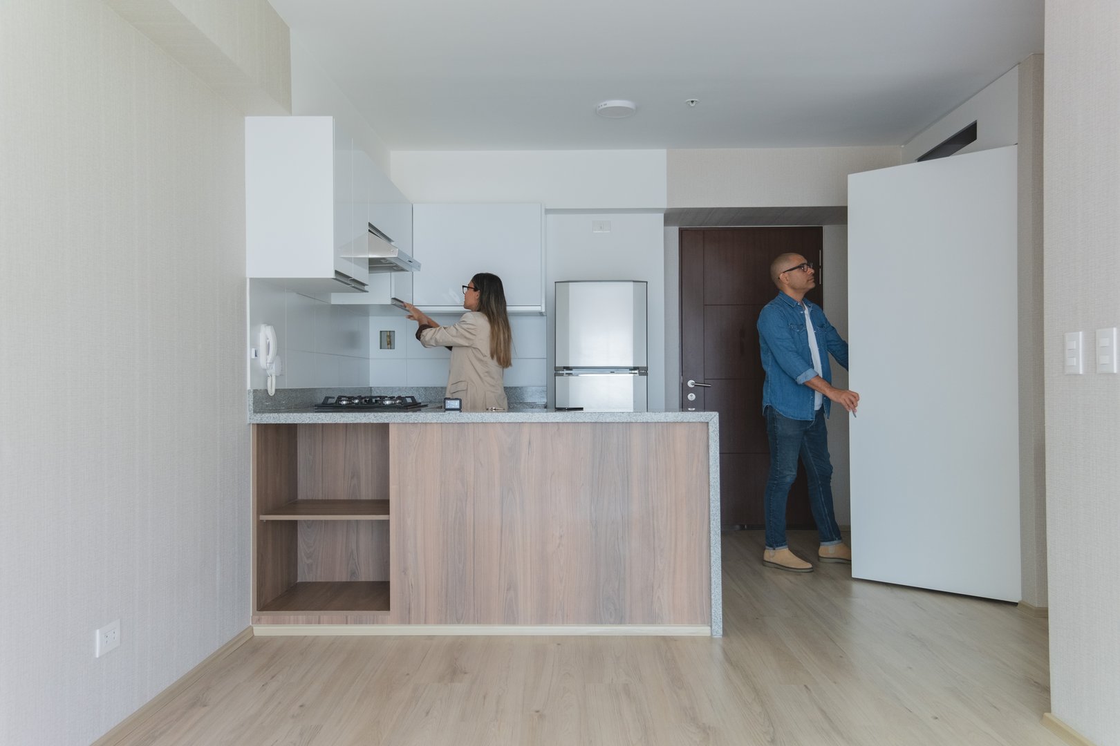 A man and a woman are standing in a kitchen. The woman is holding a remote control and the man is opening a refrigerator door. The kitchen is empty and the atmosphere is calm and peaceful