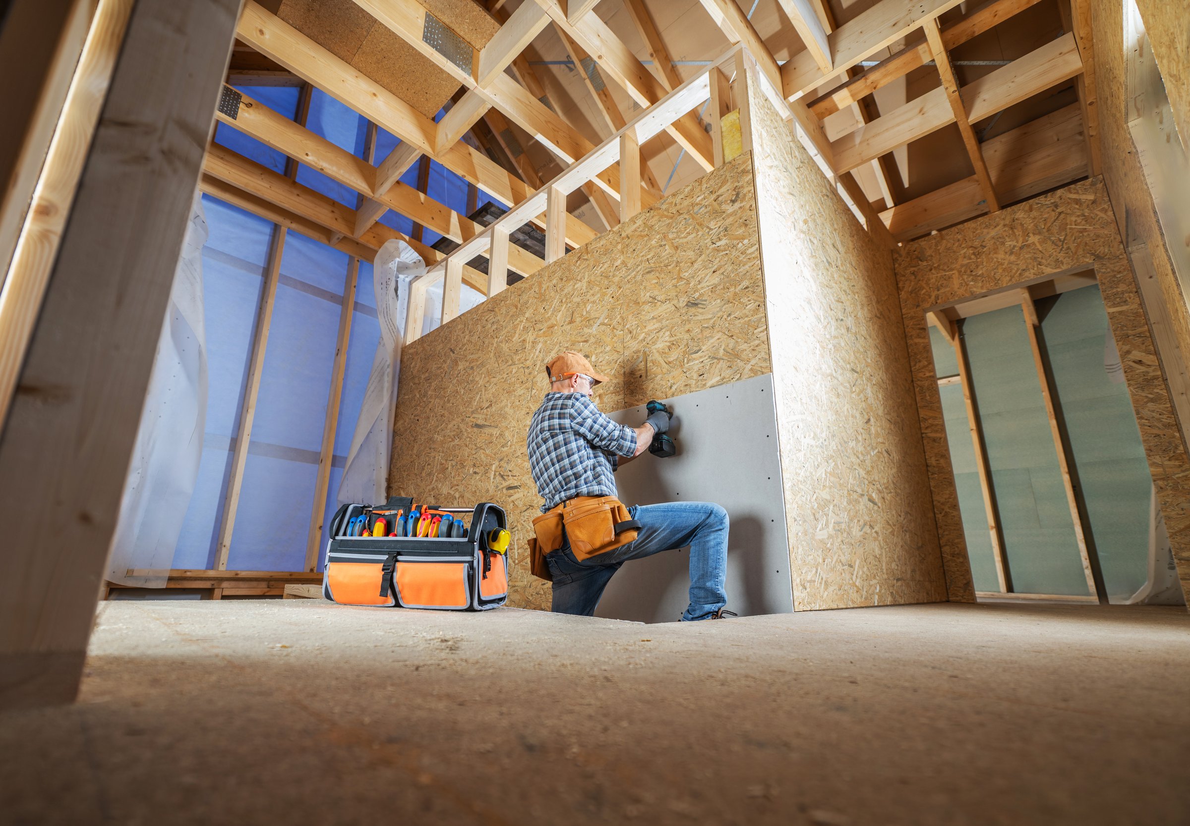 A carpenter meticulously fits drywall panels in a partially constructed room filled with wooden beams under natural light. Tools are nearby, reflecting a productive atmosphere.