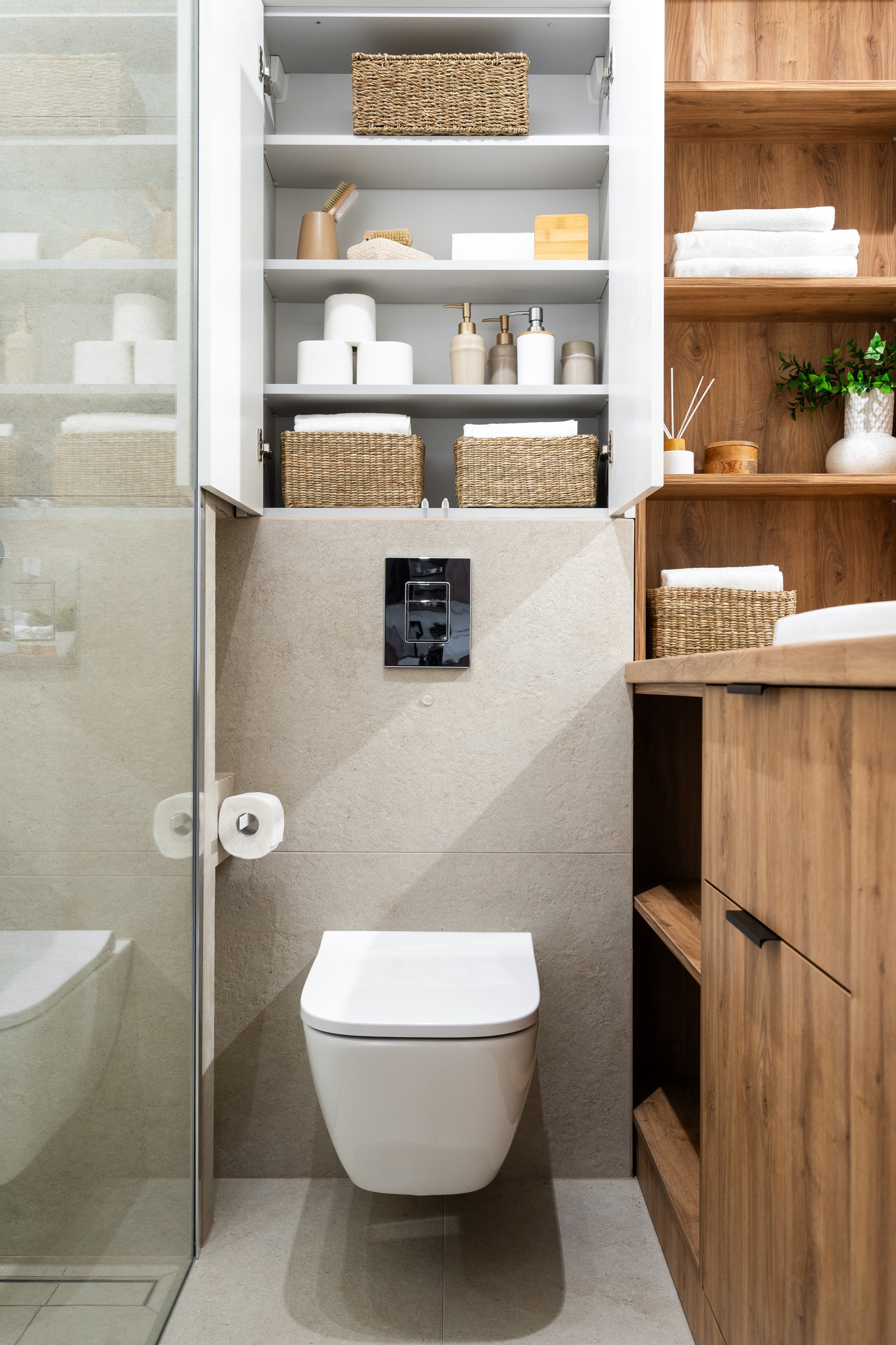 Vertical shot of bathroom interior with white wall-mounted toilet bowl, under open storage cabinet with personal accessory, toiletries, fresh towels and paper rolls