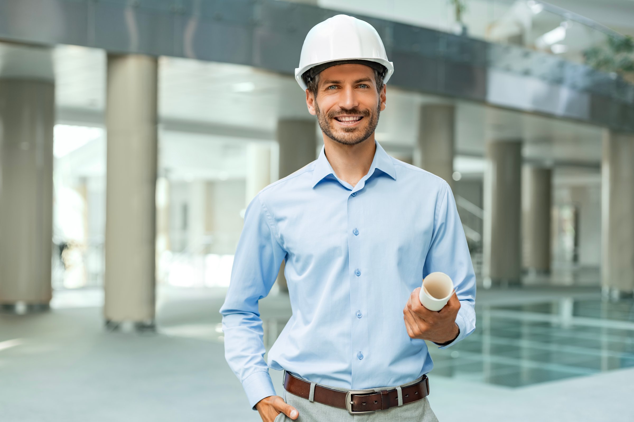 Close-up portrait of a young smiling man architect wearing a hard hat with blueprint plans looking at the camera in building under construction.
