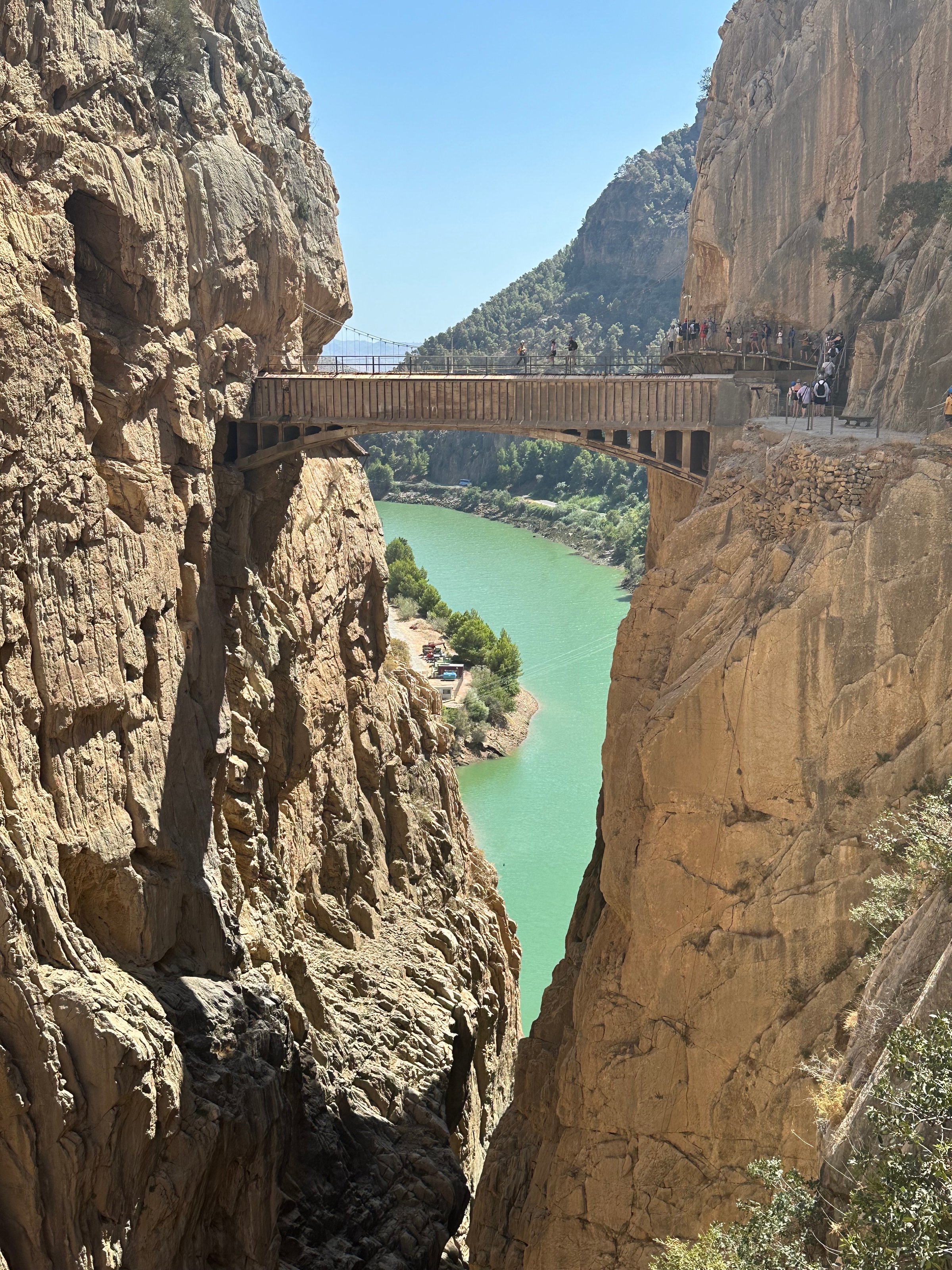 A dramatic wooden walkway clings to the steep canyon cliffs above a turquoise river, with hikers crossing the narrow path under a  bright, clear sky.