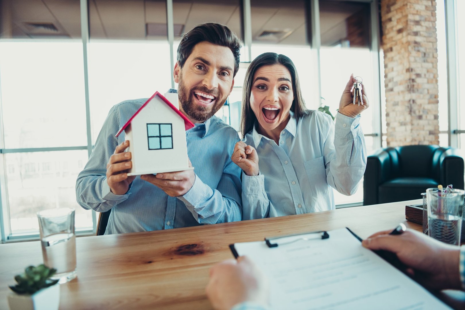 Happy couple showcasing their joy at purchasing a new property, holding house keys and a model in a modern office.