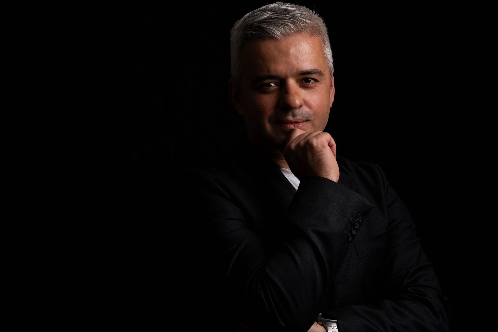 Thoughtful mature man with gray hair looking at camera, hand on chin, dramatic chiaroscuro lighting, low key studio portrait on dark background.