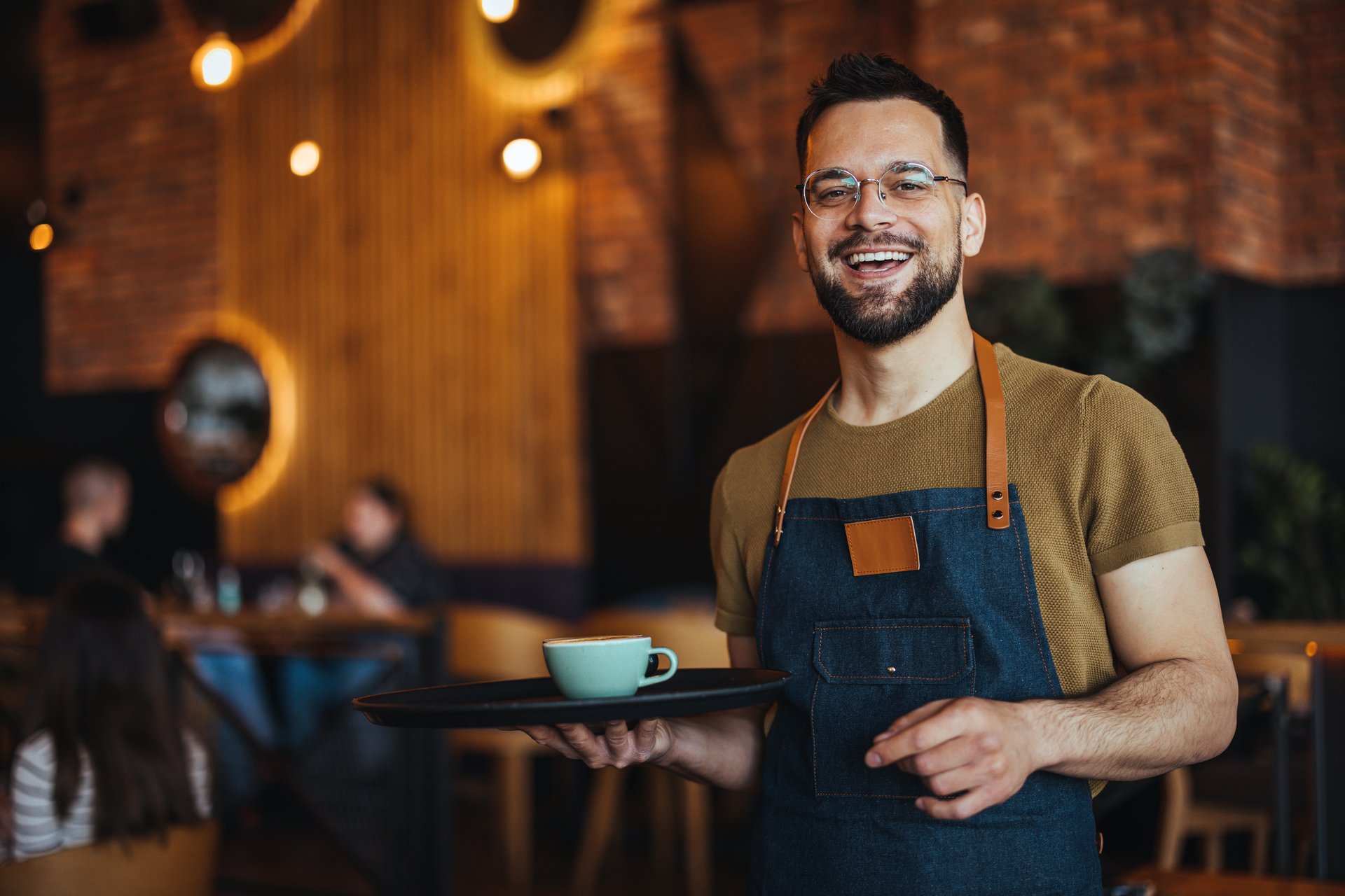 Portrait of smiling young waiter holding tray in cafe. Handsome waiter smiling at camera holding tray at the coffee shop. Profesional Waiter in Restaurant