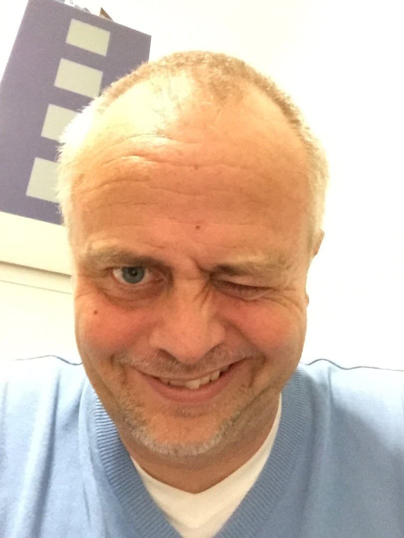 A man with short hair winks at the camera, wearing a light blue sweater, smiling in a close-up selfie.