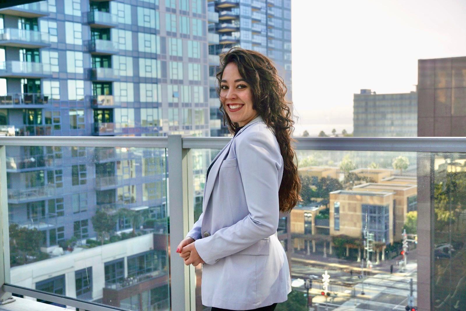 Woman in a light blazer smiling on a balcony, with a cityscape backdrop of modern buildings and a street below.