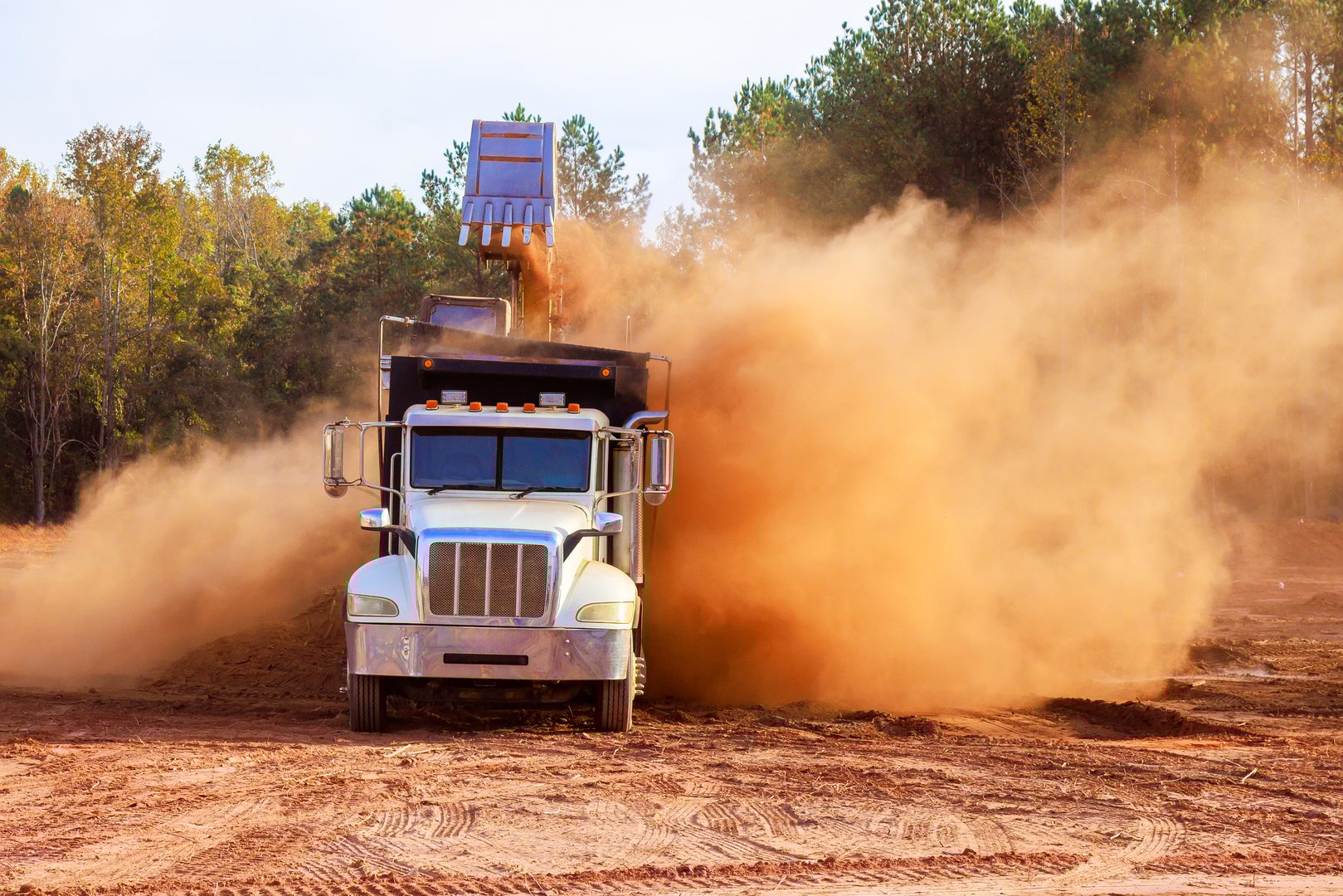 Construction truck is unloading materials at job site, sending cloud of dust into air is construction area surrounded by during land removal