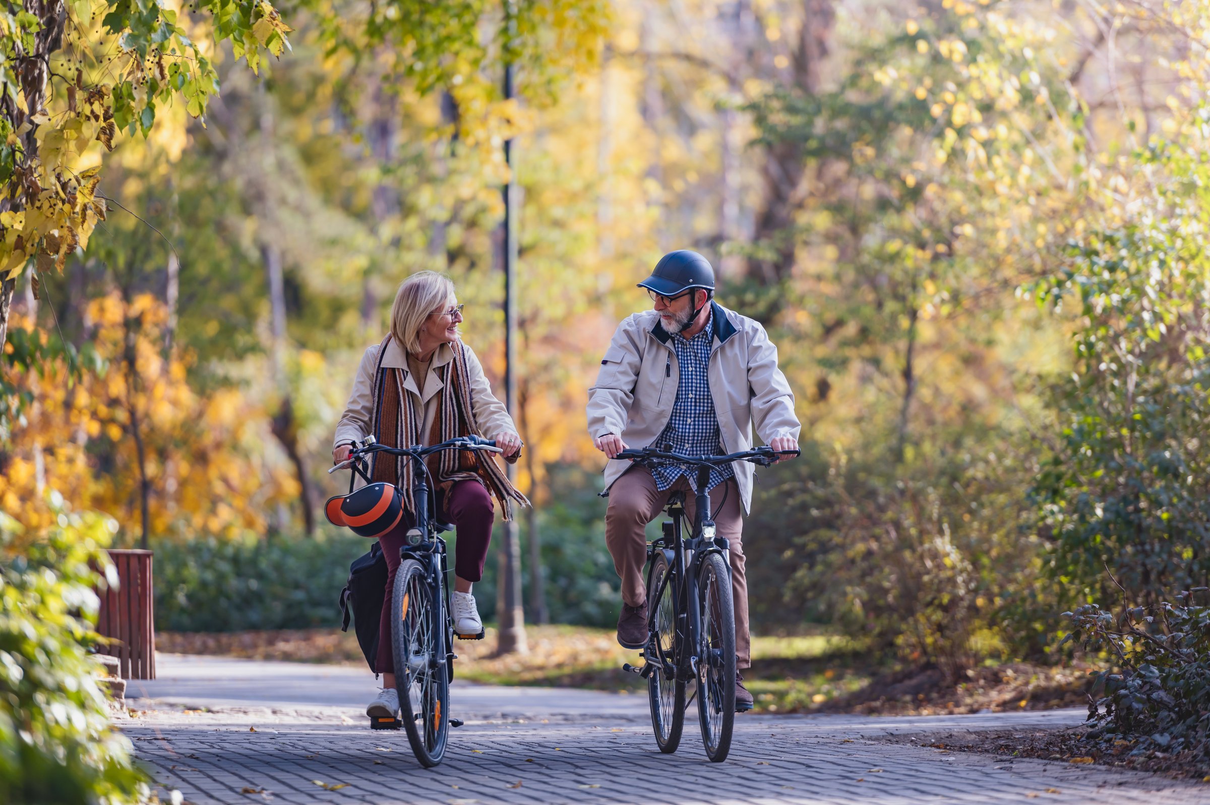 Senior couple riding bicycles together on a paved path in an autumn park, smiling and enjoying a peaceful outdoor activity surrounded by colorful trees.