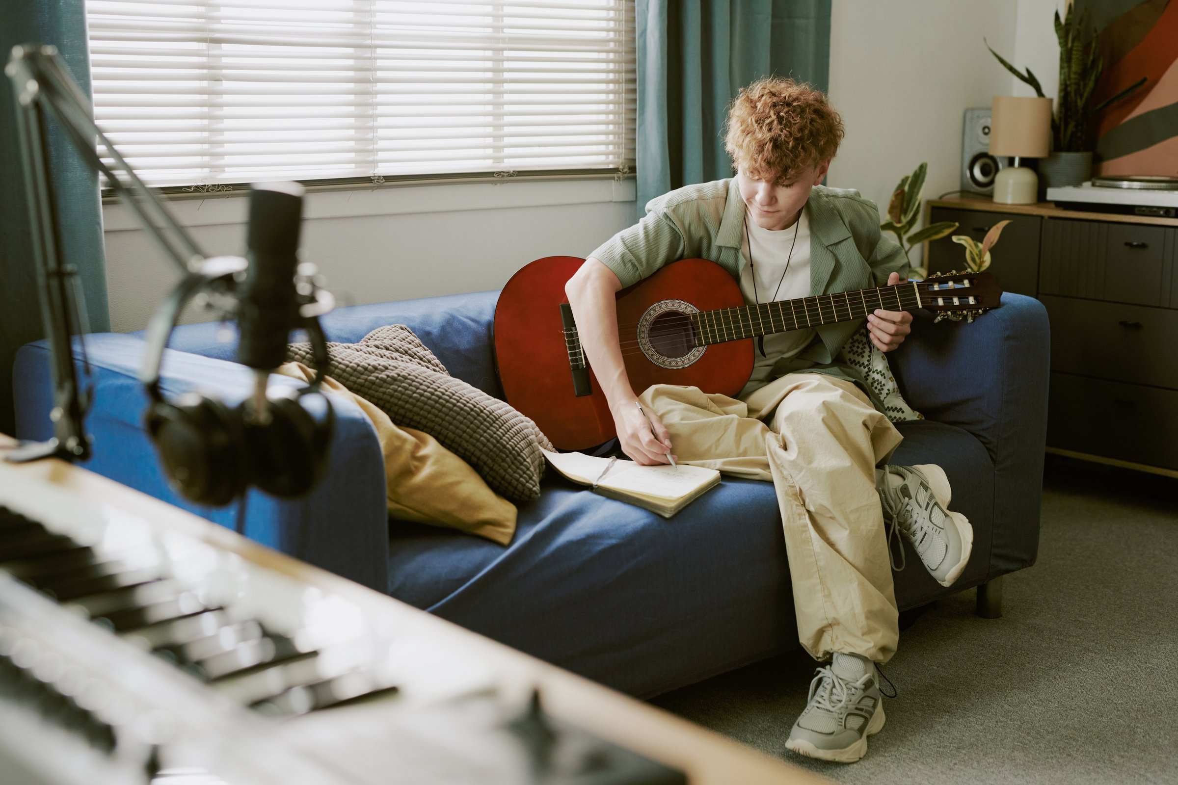 Caucasian teenage boy sitting on sofa playing acoustic guitar and writing music in notebook, recording audio with microphone and headphones, digital piano visible in foreground