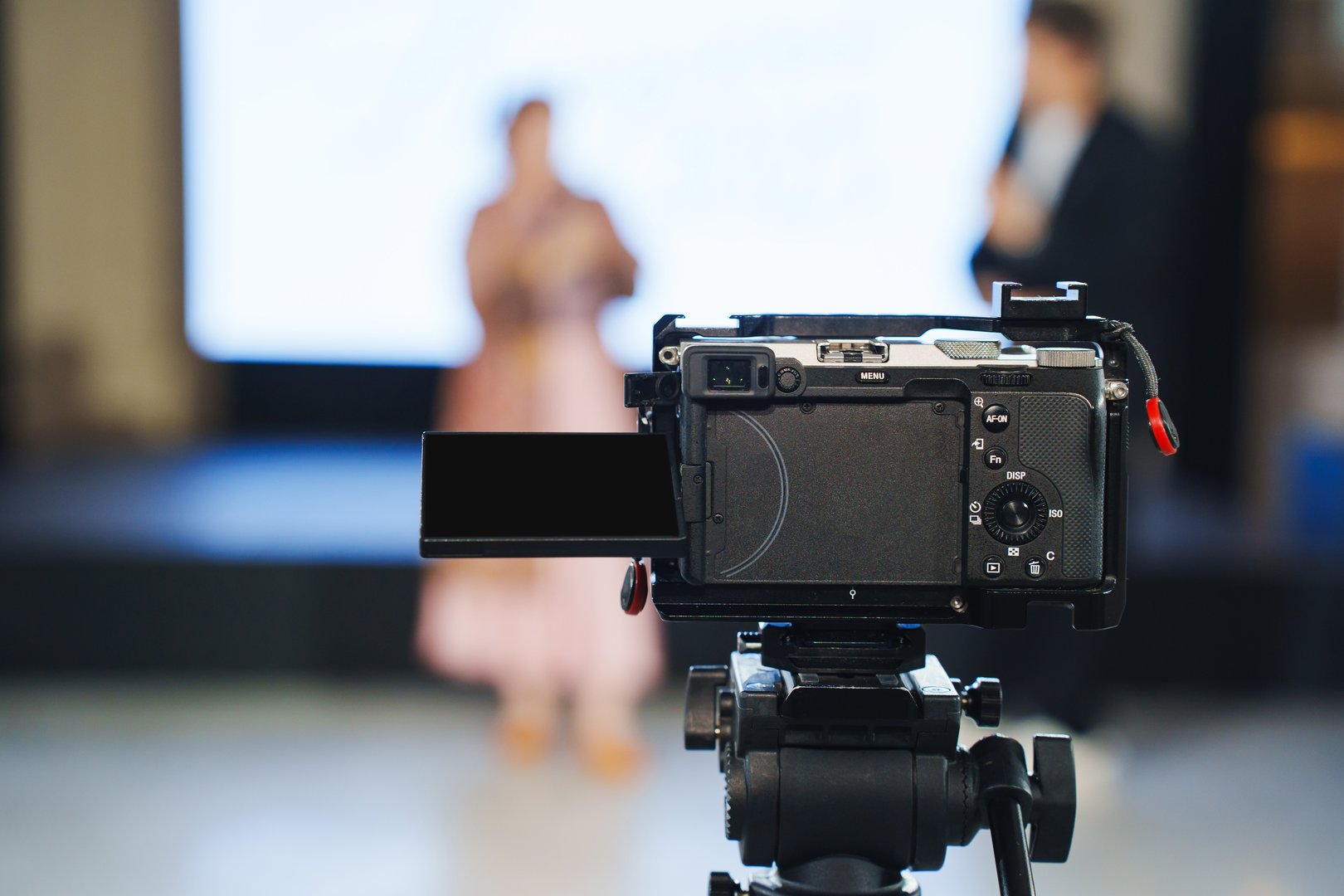 A camera is positioned on a tripod, focusing on two presenters in a large indoor venue. The background features a screen displaying digital content. The atmosphere is professional.