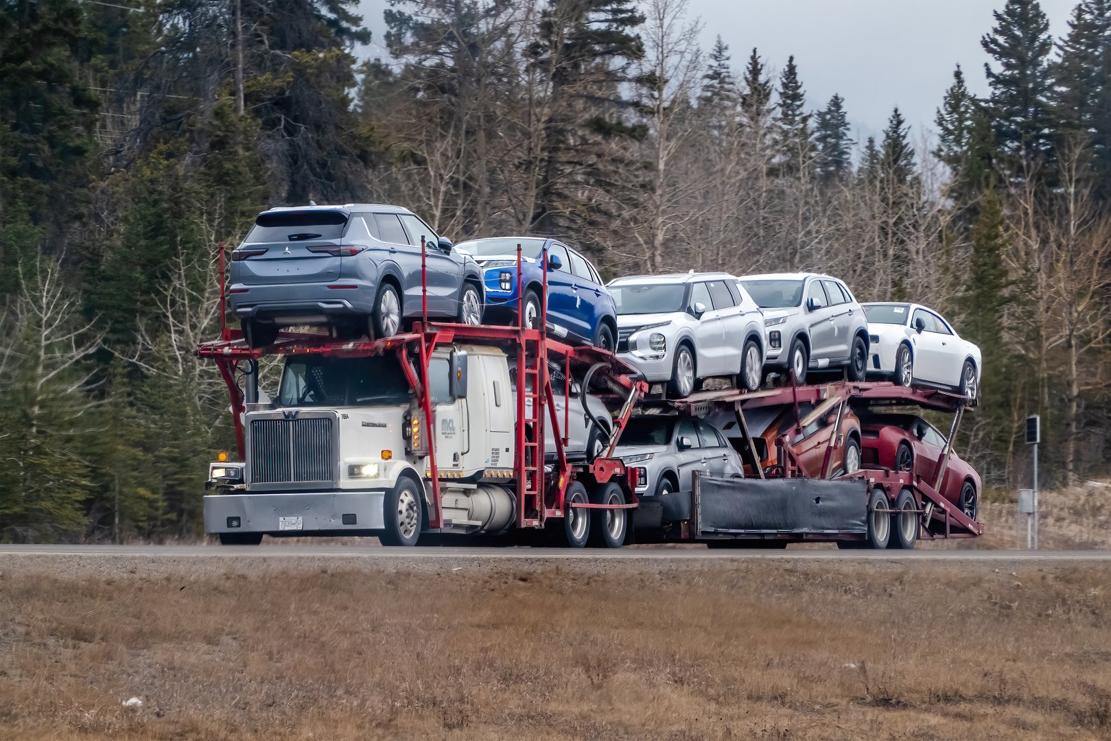 Calgary, Alberta, Canada. Apr 5, 2025. Multi-level car hauler transports several new vehicles along a tree-lined highway, showcasing automotive logistics in action.