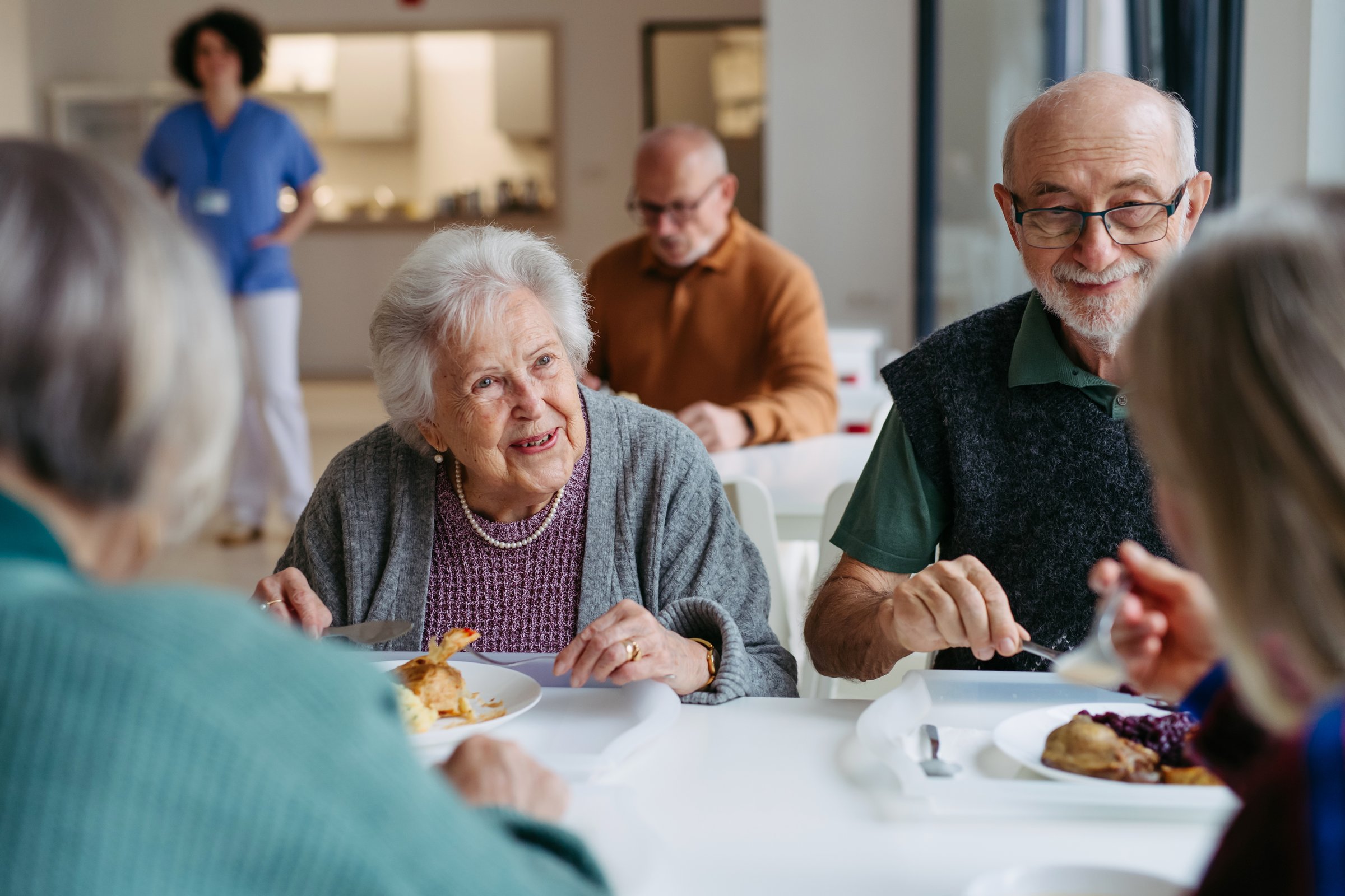 Group of elderly people having lunch with supportive caregiver in community center cafeteria.