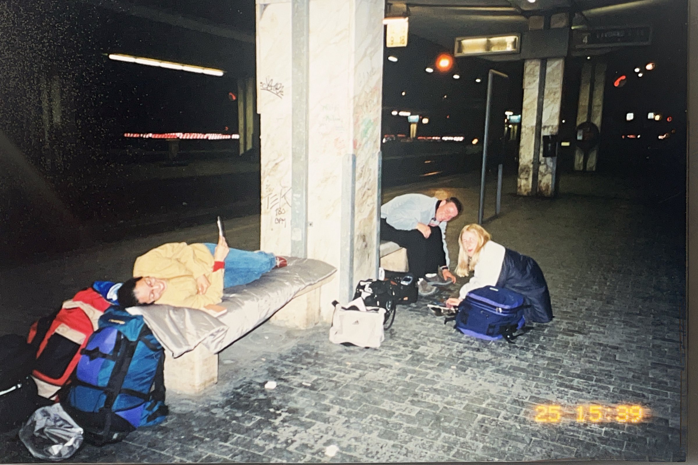 Three people with backpacks resting at a bus station, one lying down on a bench and two seated on the ground with bags.