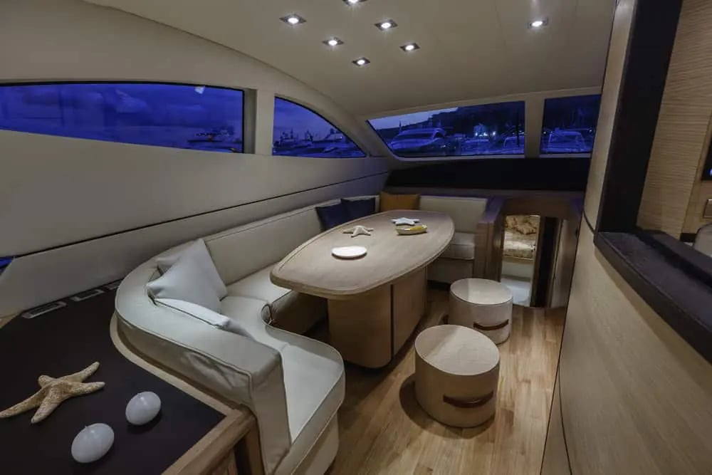A luxury yacht interior lounge with elegant cream-colored L-shaped seating, wooden flooring, and large windows offering stunning views. The space features modern design with ambient lighting and sophisticated decor.