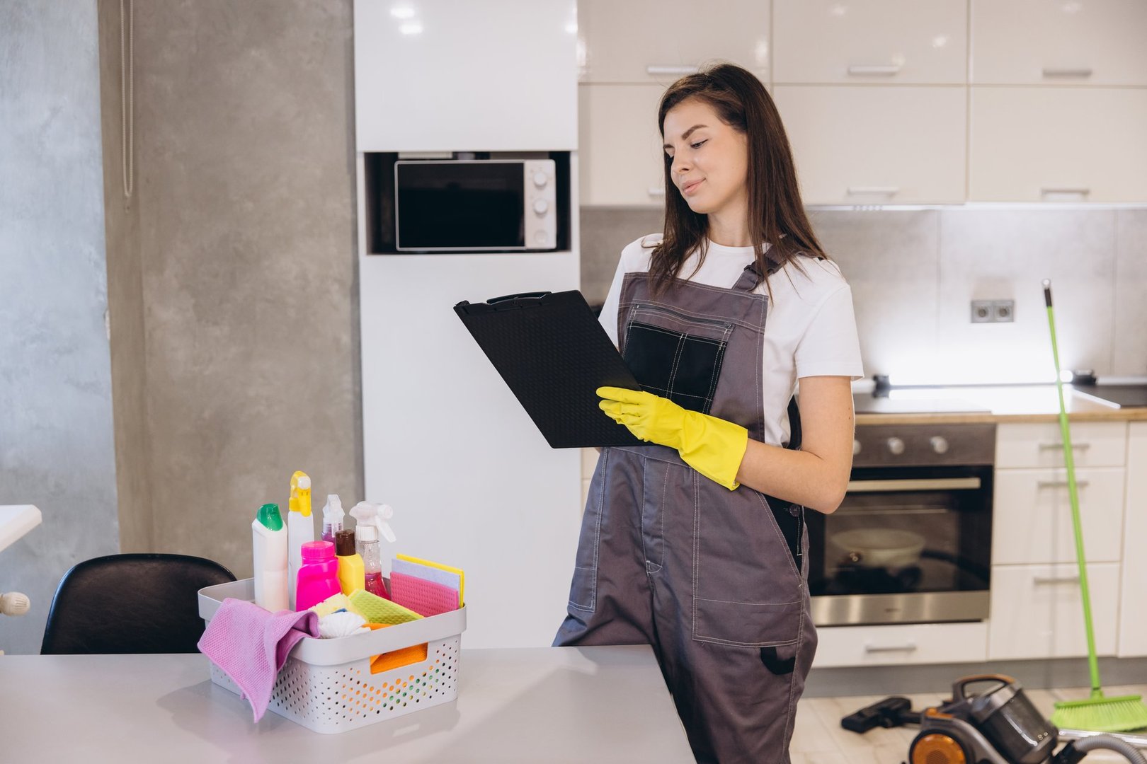 Professional cleaning lady wearing yellow gloves and grey uniform checking the list of cleaning products standing in a modern kitchen