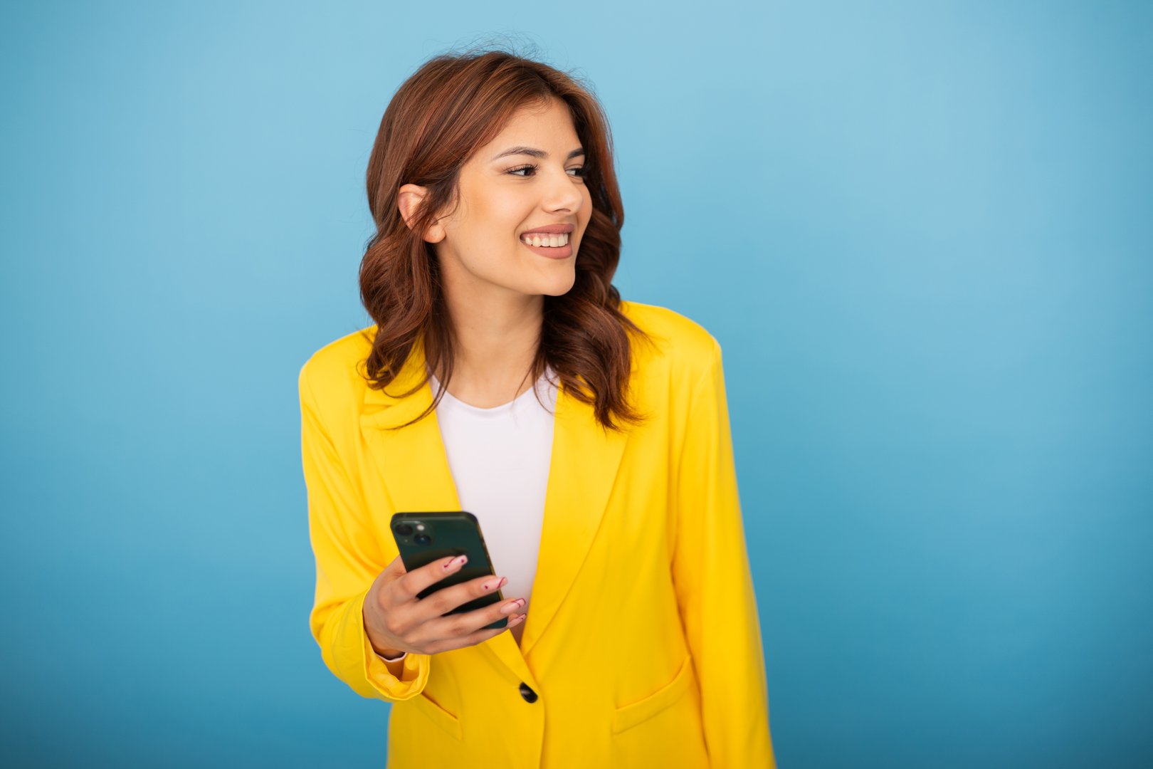 Cheerful businesswoman using mobile phone, looking away and smiling on blue background