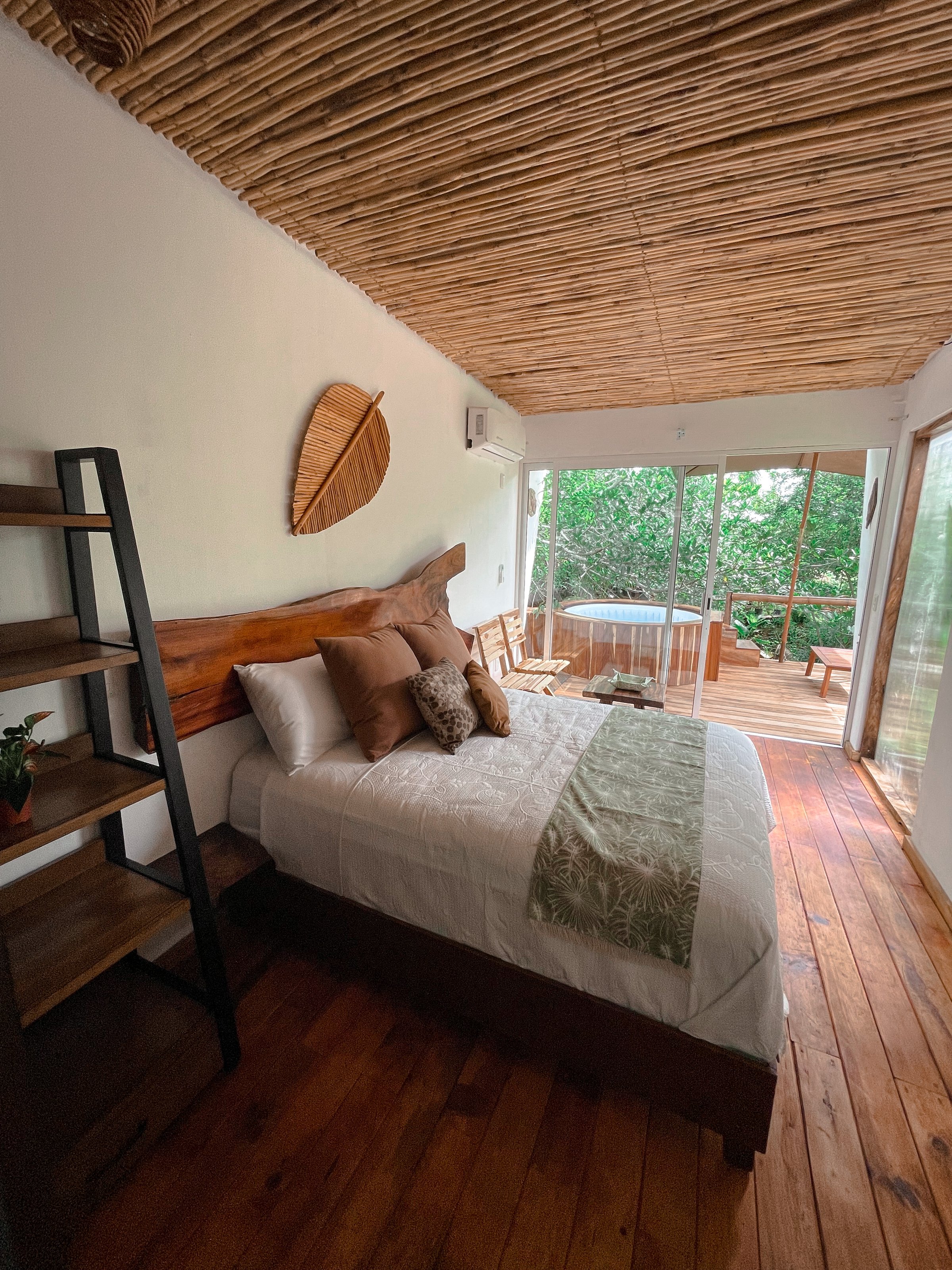 A cozy bedroom with a wooden ceiling, bed, and floor. Large windows open to a deck and trees outside.