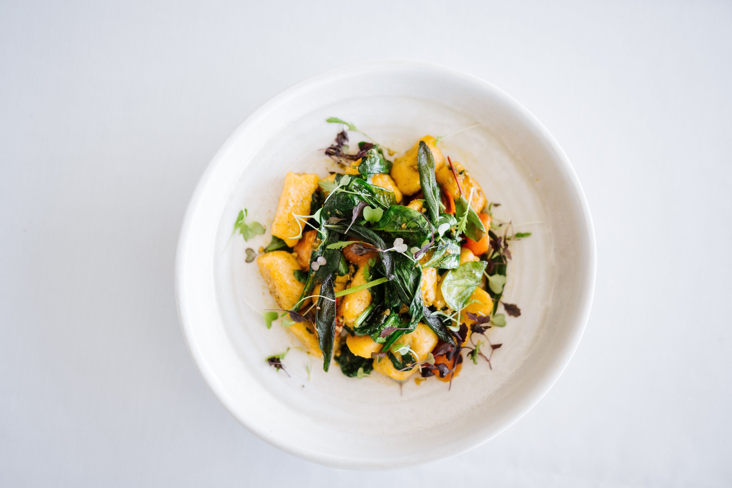 A top view of a gourmet dish with gnocchi and fresh greens on a white plate, set against a white background.