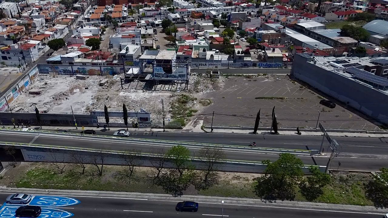 Aerial view of an urban area with roads, parking lots, and residential buildings. Sparse trees line the streets.