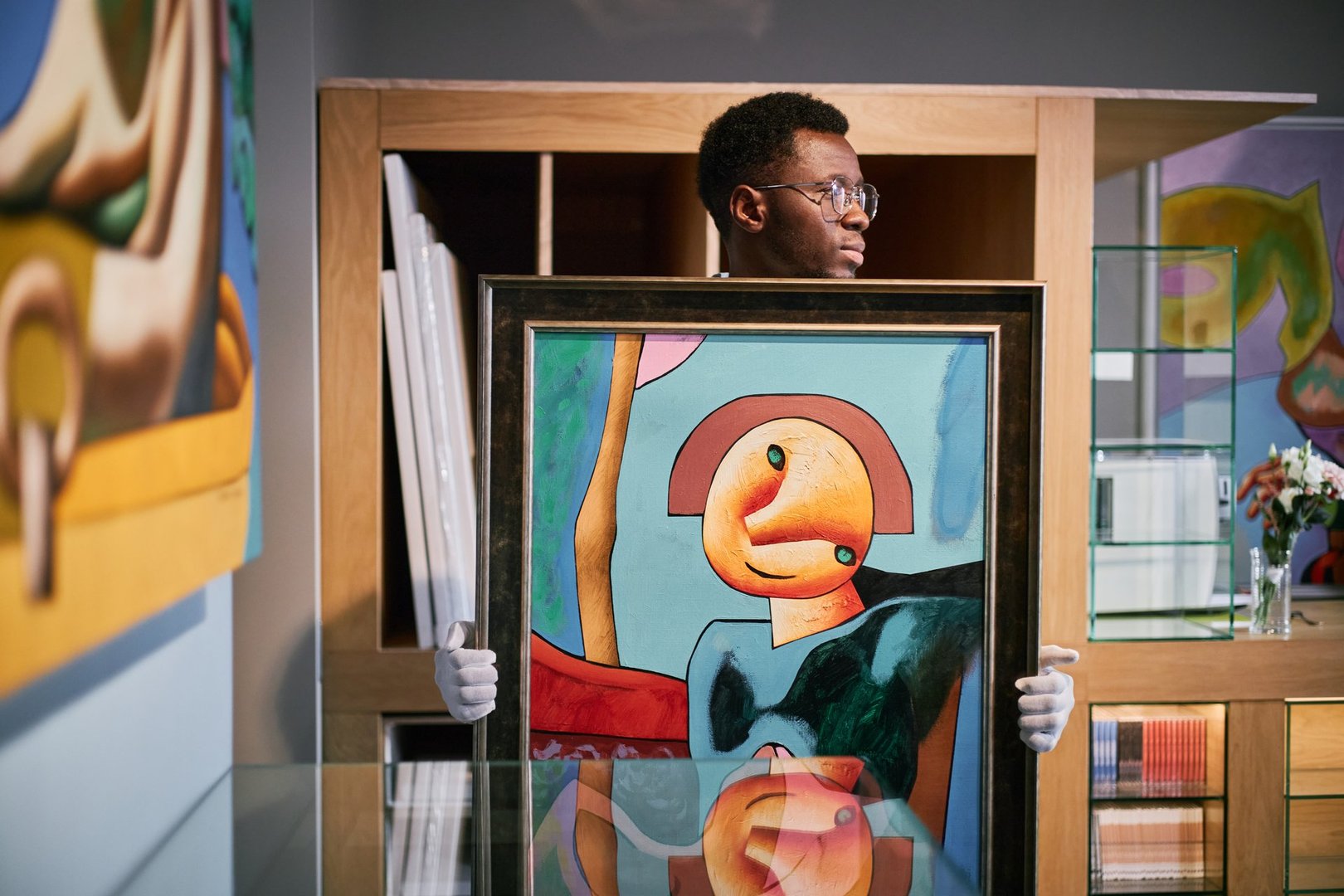 Black person holding framed modern abstract painting in art gallery with other artworks displayed around and reflection visible on the glass surface