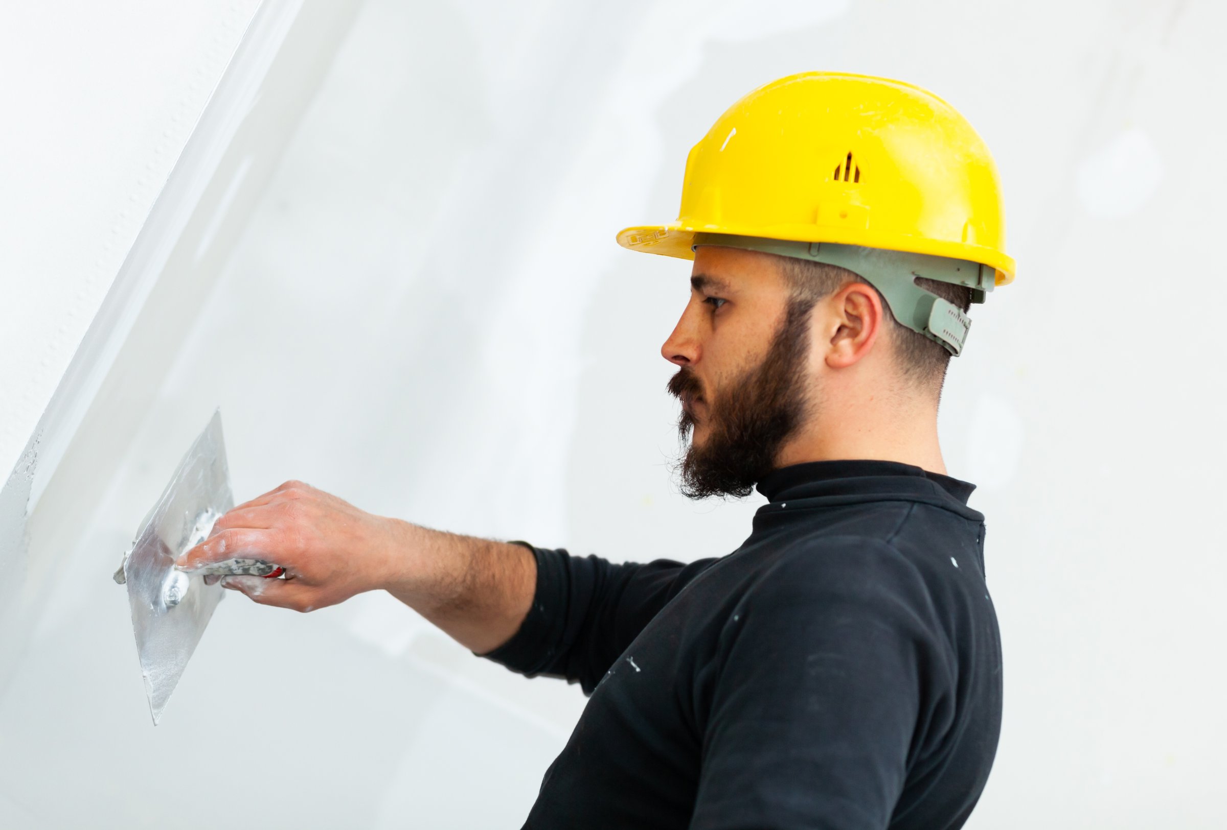 Construction worker wearing yellow hard hat smoothing white wall with putty knife