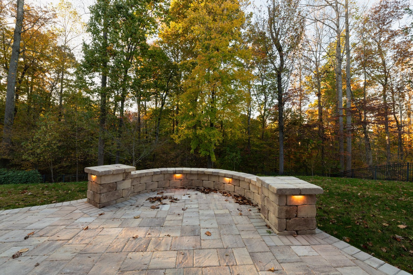 Picturesque backyard view in autumn season with patio pavers and stone wall, autumn leaves, and colorful woods in the background.