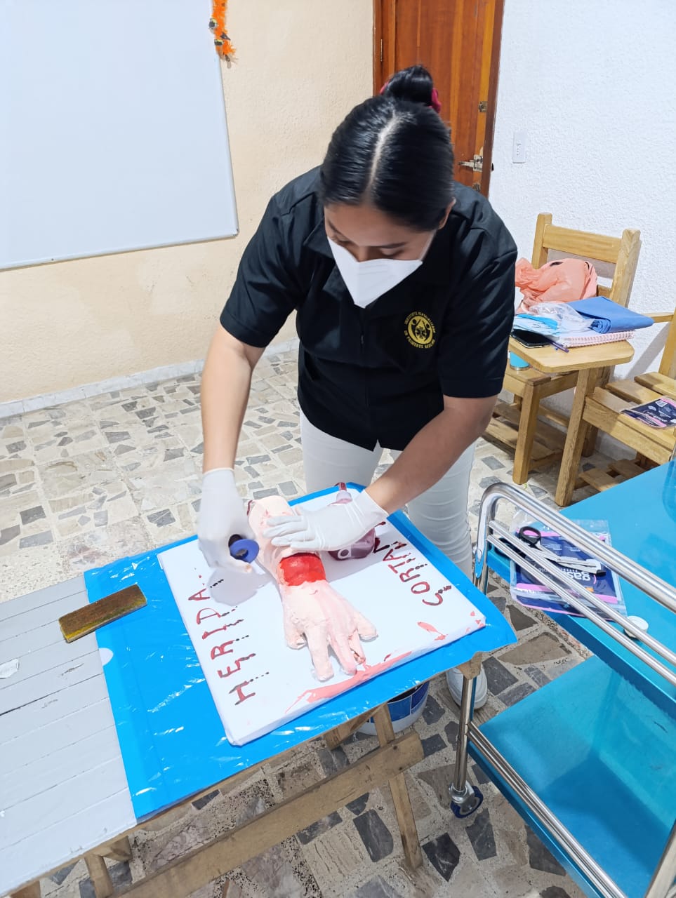 A person in a mask applies a bandage to a realistic training model of an injured arm on a table in a medical setting.