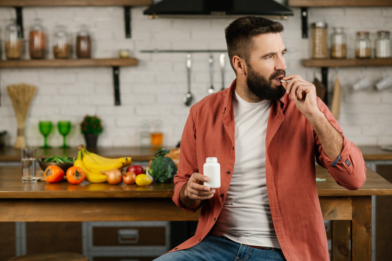 Young attractive man sitting at the kitchen table and drinking dietary supplements, looking away. Healthy lifestyle concept.
