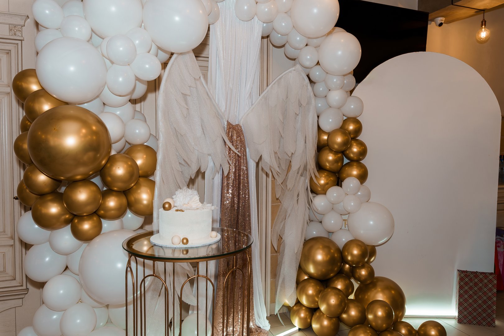 A beautifully styled first birthday party features a stunning white and gold balloon arch framing an angel-winged setup with a small cake, capturing joyous celebration vibes