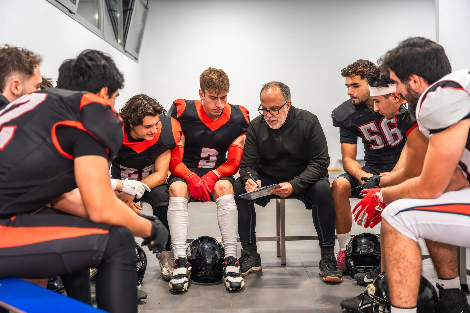 American football coach discussing game plan with young men players, wearing uniforms and protective gear, sitting on benches in the locker room, emphasizing teamwork and communication