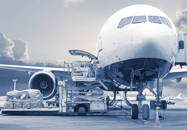 Cargo being loaded onto a large commercial airplane at an airport, with equipment and pallets in foreground.