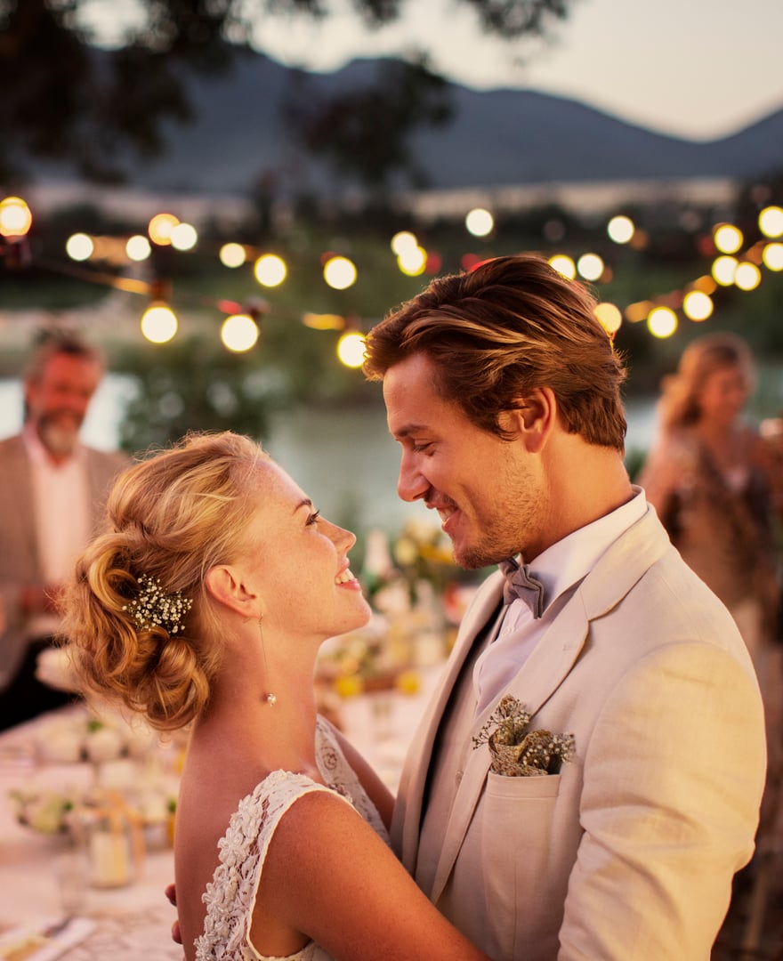 A bride and groom smiling at each other during an outdoor evening wedding with string lights and guests in the background.