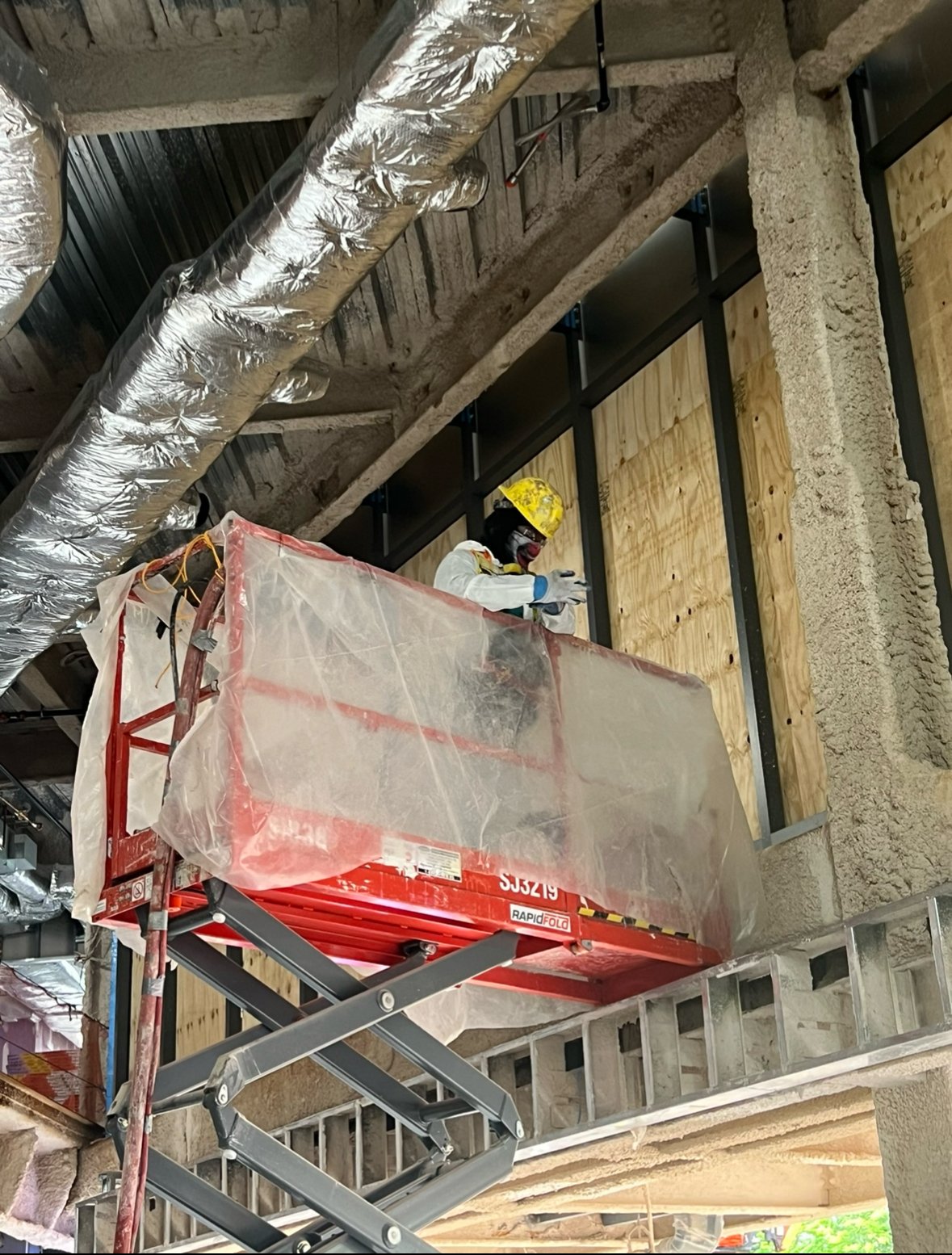 Worker on scissor lift wearing safety gear, applying material to ceiling near ventilation ducts in a construction site.