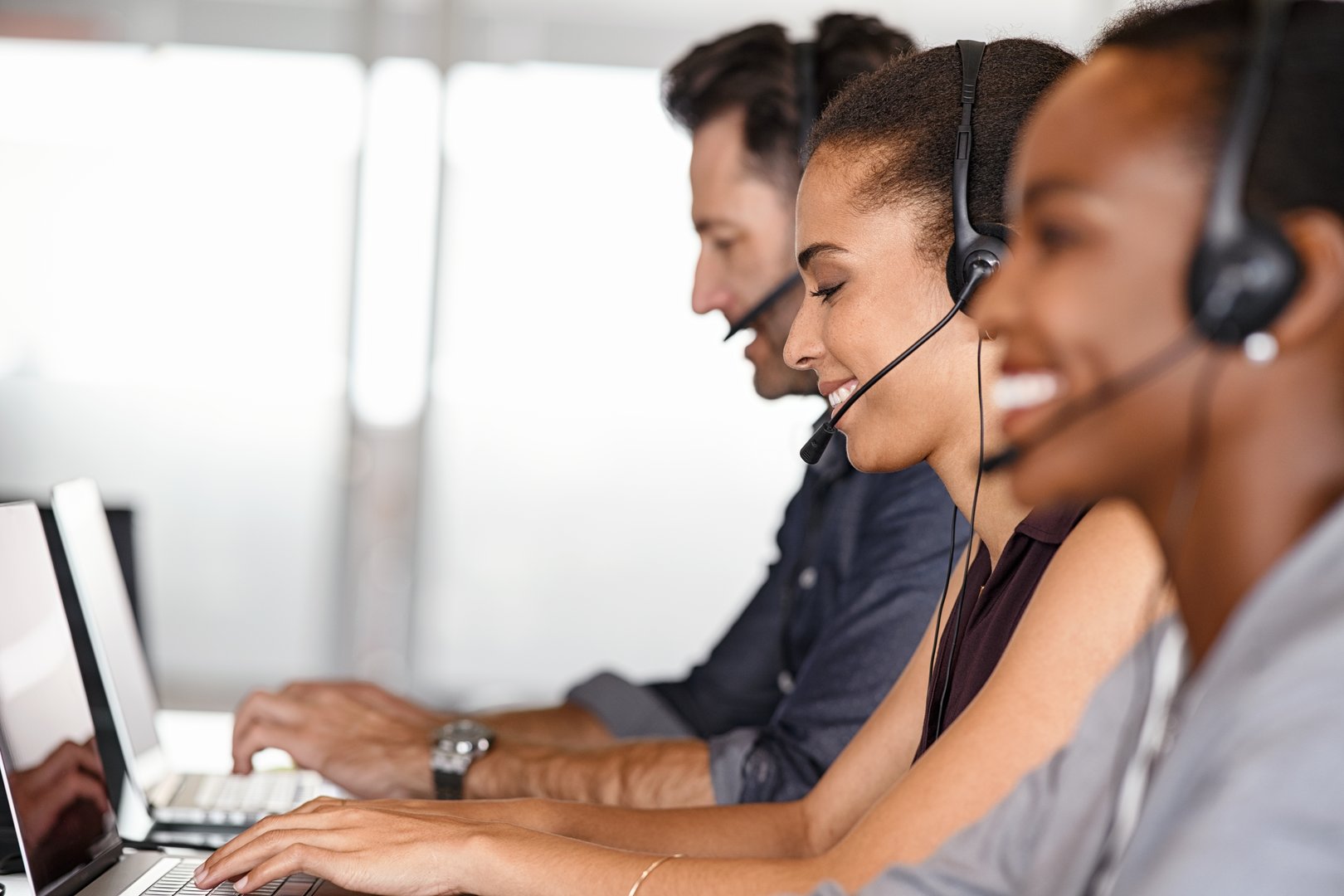 Smiling multiethnic people with headsets using computer and smiling while working in office. Young man and woman operators talking on headset with clients. Group of telemarketing customer service team working while sitting in a row.