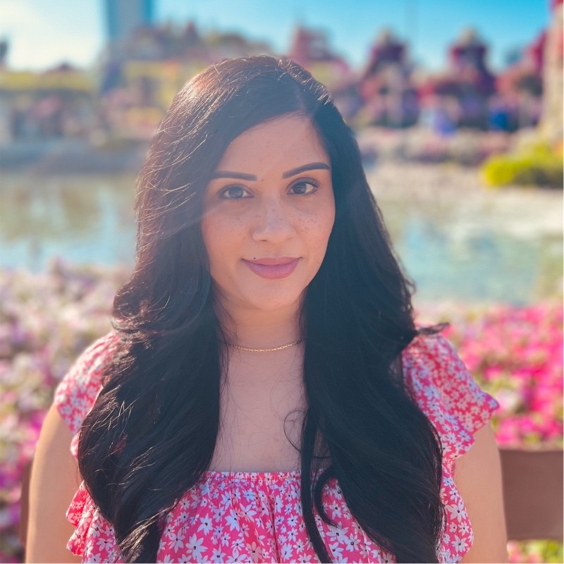 Woman with long dark hair and a floral dress stands outdoors with a blurred scenic background.