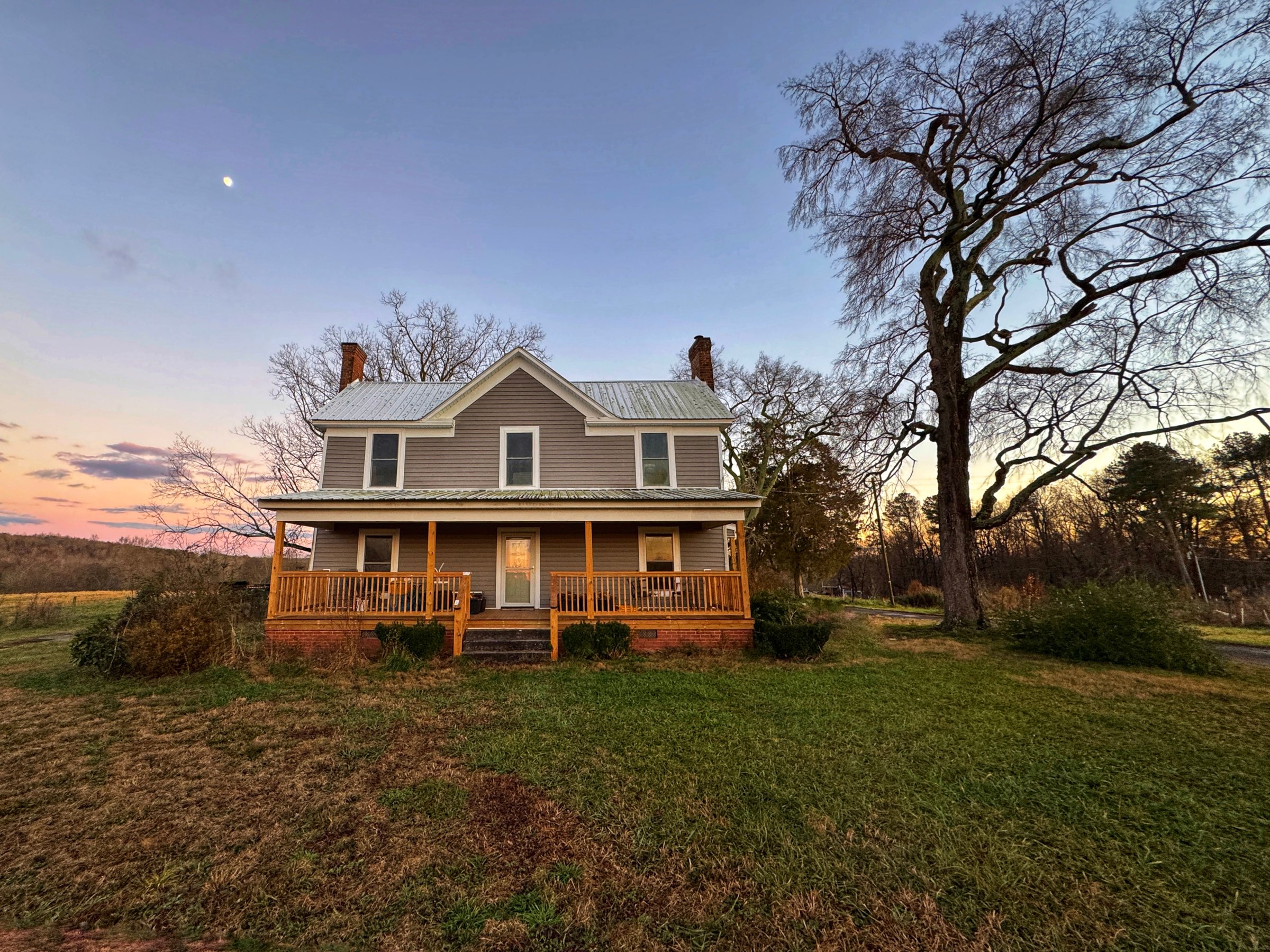 A wind swept, craggy tree next to grey farmhouse built in 1920 with a new and renovated wooden porch. The moon rising overhead