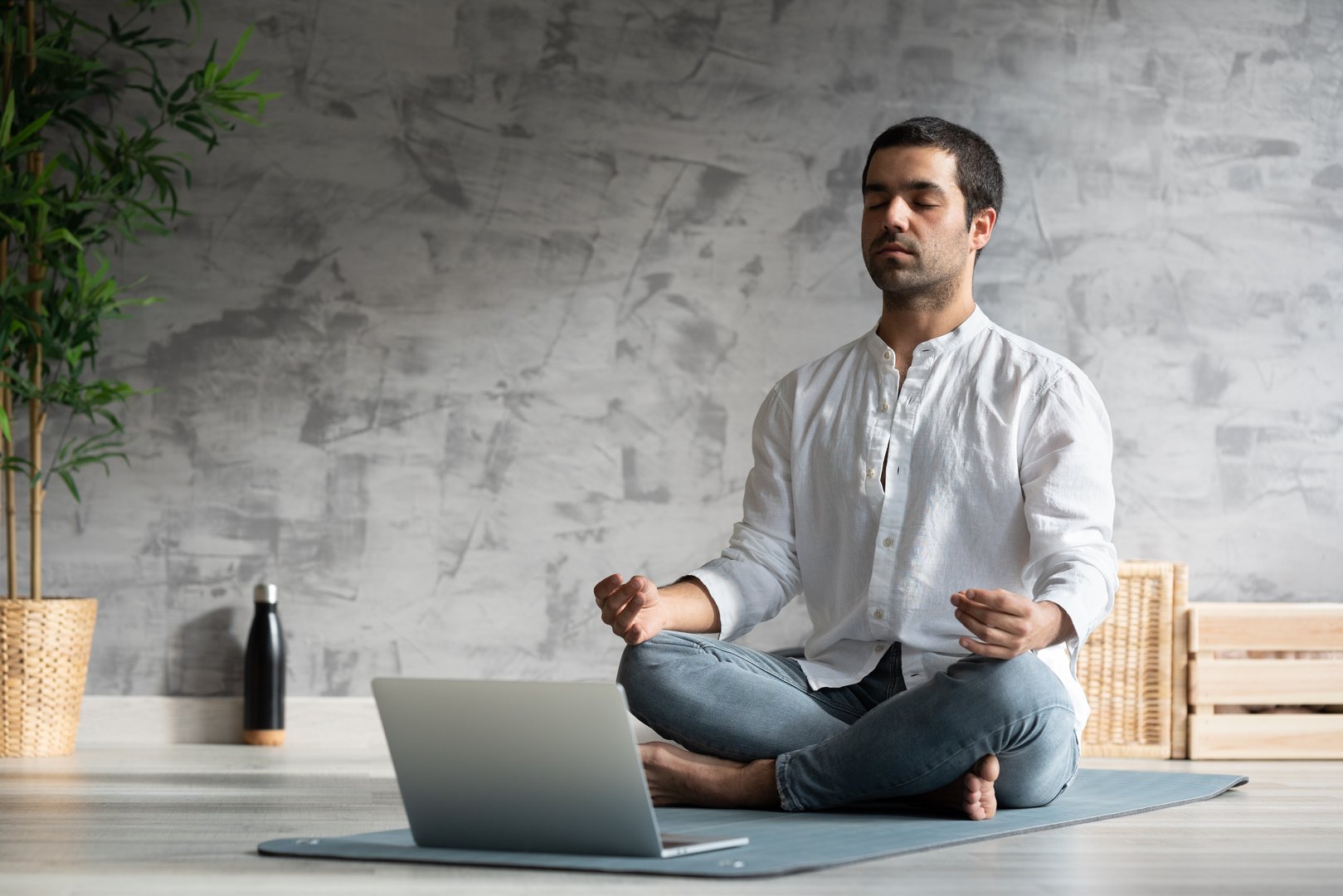 Hispanic yoga teacher meditating in front of laptop indoors. Student following meditation tutorial sitting on a mat