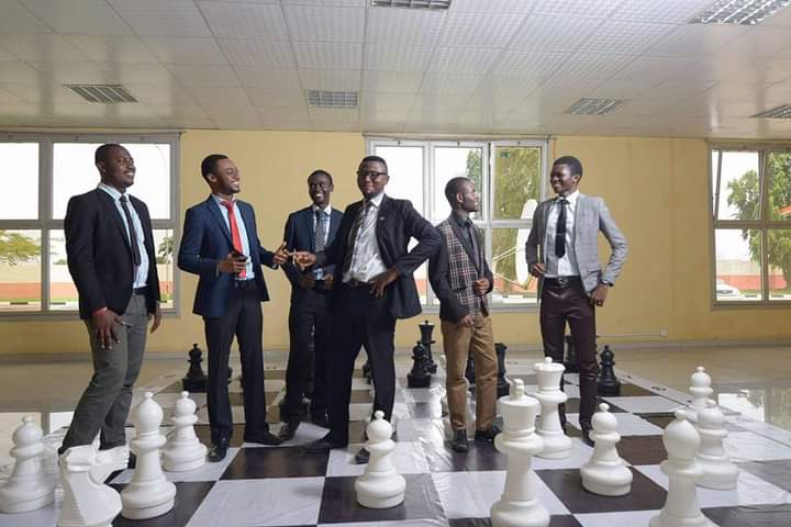 Six men in suits stand on a large indoor chessboard with oversized chess pieces, smiling and interacting.