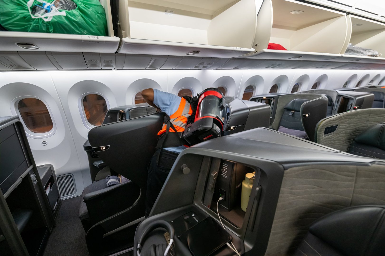 A Middle-Eastern male airline crew member in a bright orange safety vest is meticulously vacuuming the airplane cabin. The modern aircraft interior features comfortable, spacious seats and overhead compartments.